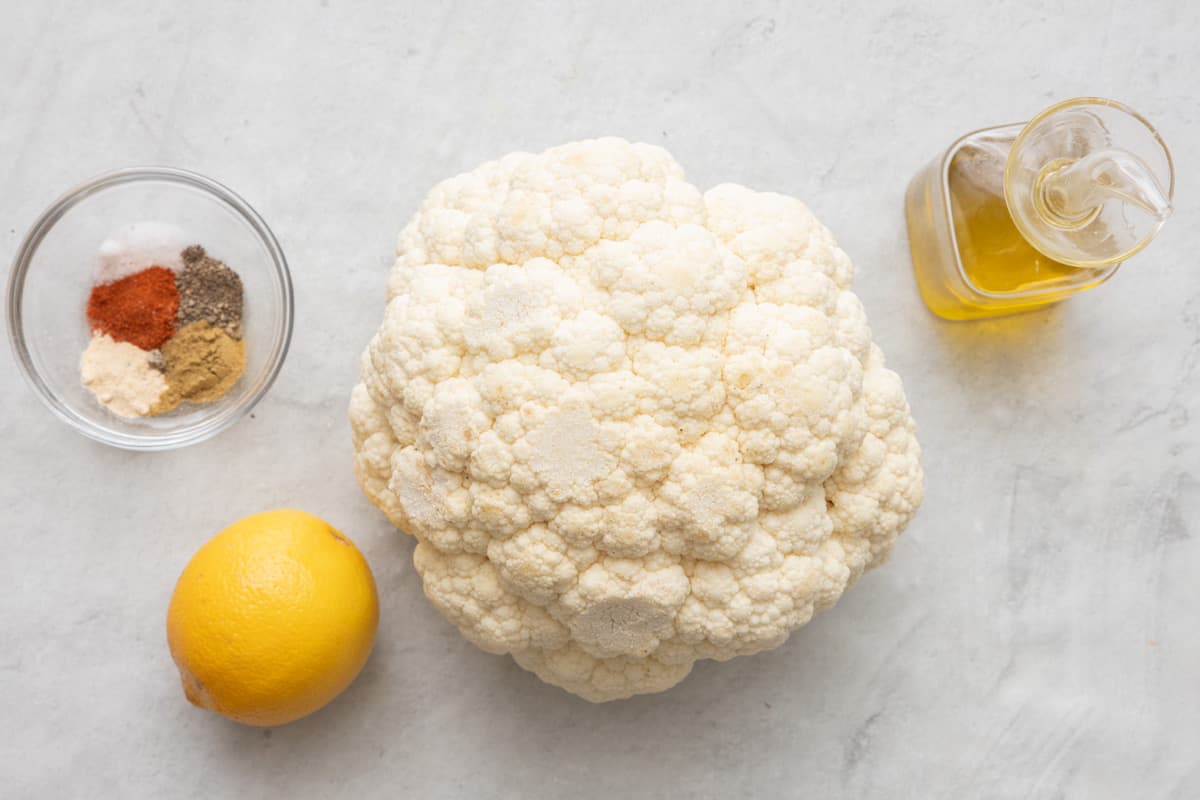 Ingredients for recipe: spices in small bowl, a lemon, whole head of cauliflower, and oil.
