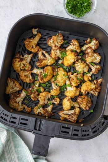 Brown and crispy cauliflower florets in an air fryer basket garnished with fresh parsley.