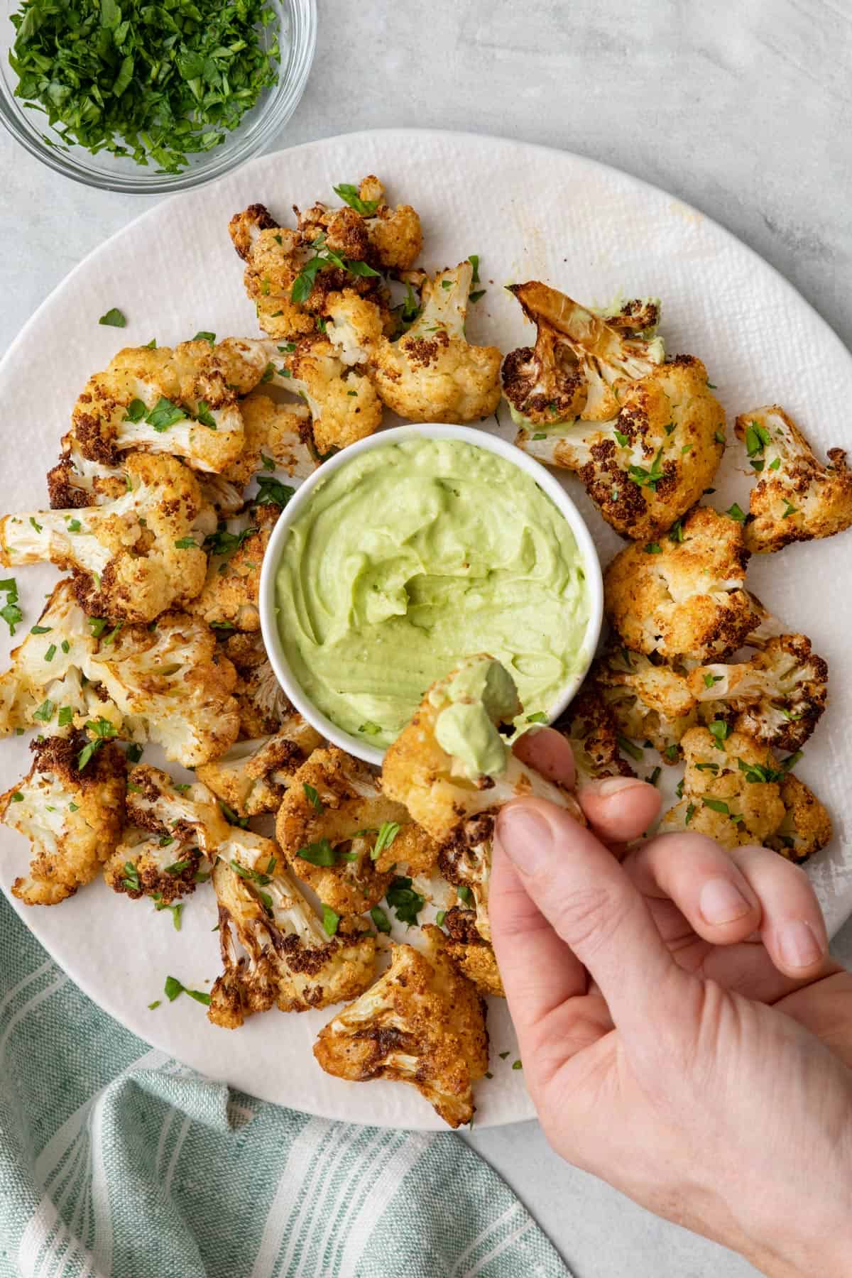 Hand holding a piece of cauliflower dipped in thick avocado crema over a plate of more bite sized pieces.