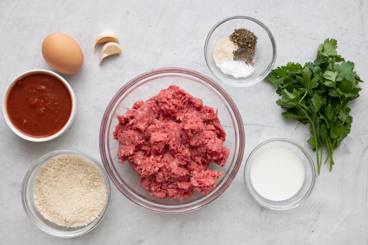 Ingredients for recipe in individual bowls and before prepped: marinara sauce, an egg, garlic cloves, breadcrumbs, seasonings, milk, and fresh parsley.
