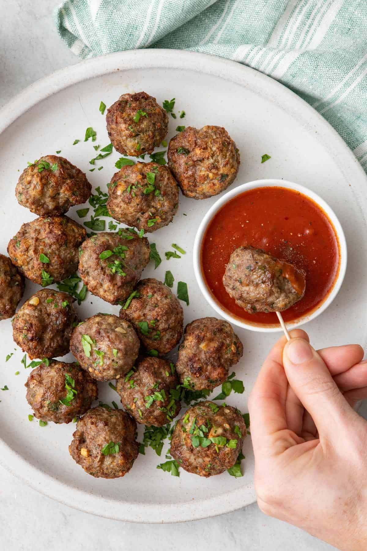 Dipping a meatball on a toothpick into marinara sauce with more meatballs on plate.