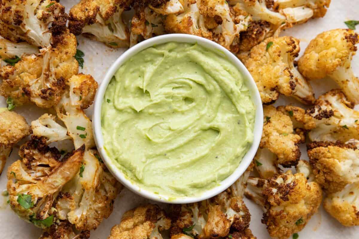 A small bowl of creamy avocado dip surrounded by roasted cauliflower florets.