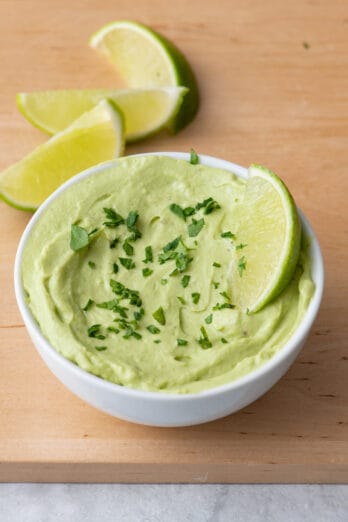 Avocado crema in serving dish garnished with chopped herbs and lime wedges.