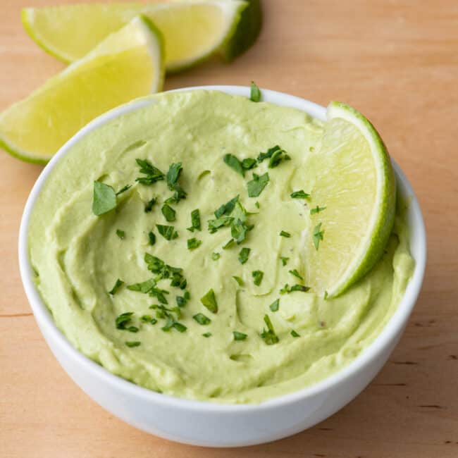 Avocado crema in serving dish garnished with chopped herbs and lime wedges.