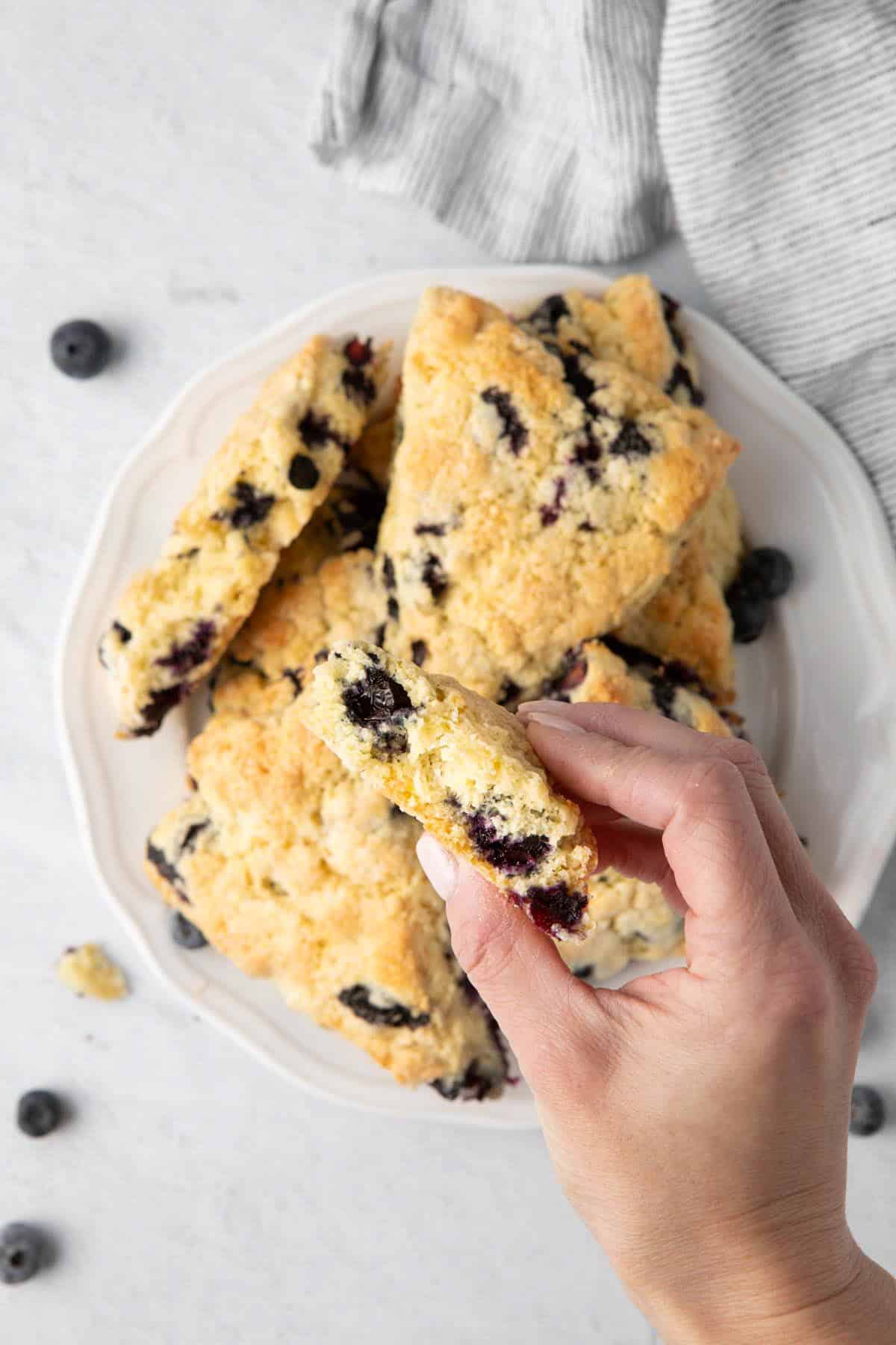 Overlapping scones on plate, with one on plate ripped in half and a hand holding the other close to show texture inside.