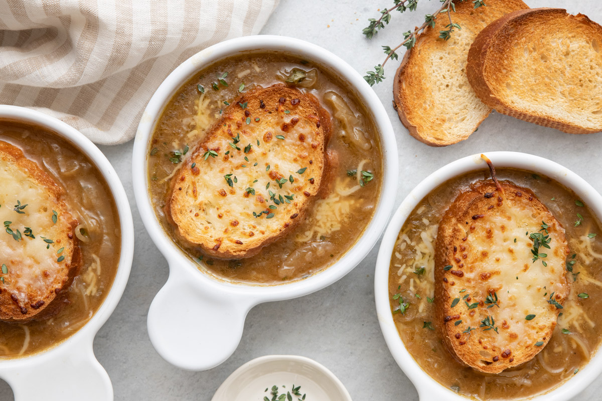 3 bowls of French onion soup with toasty sliced bread and melty cheese on top and a few extra toasted bread nearby.