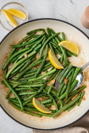 Green beans in pan after cooked garnished with lemon wedges and a serving spoon dipped in.