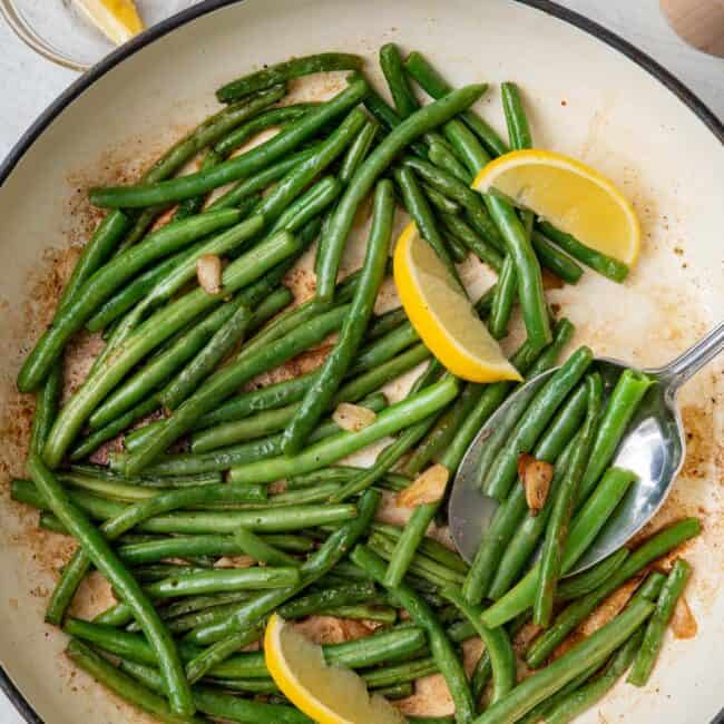 Green beans in pan after cooked garnished with lemon wedges and a serving spoon dipped in.