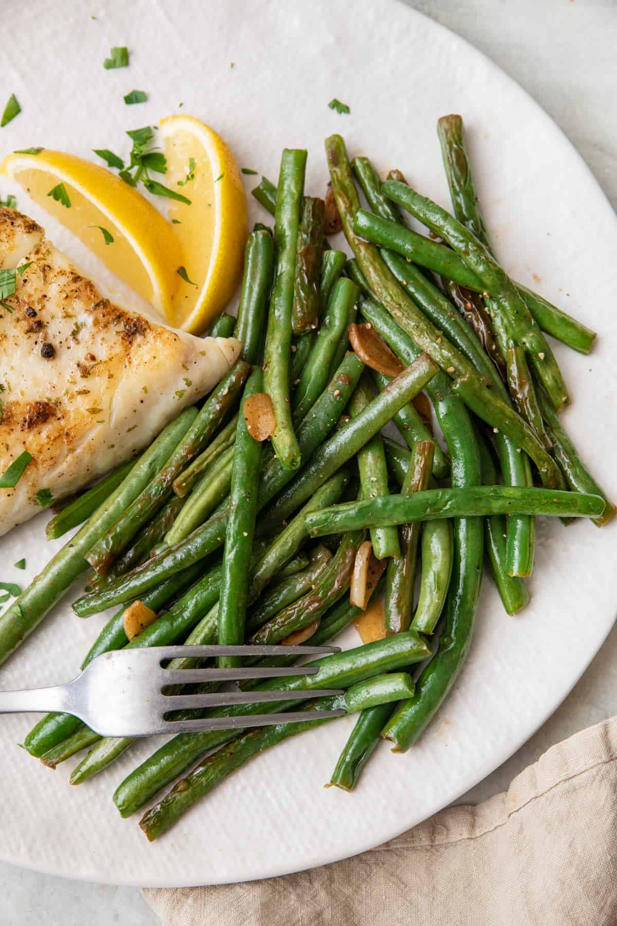 Sauteed green beans on a plate with a fork punctured into a few next to a piece of cooked fish and lemon wedges.