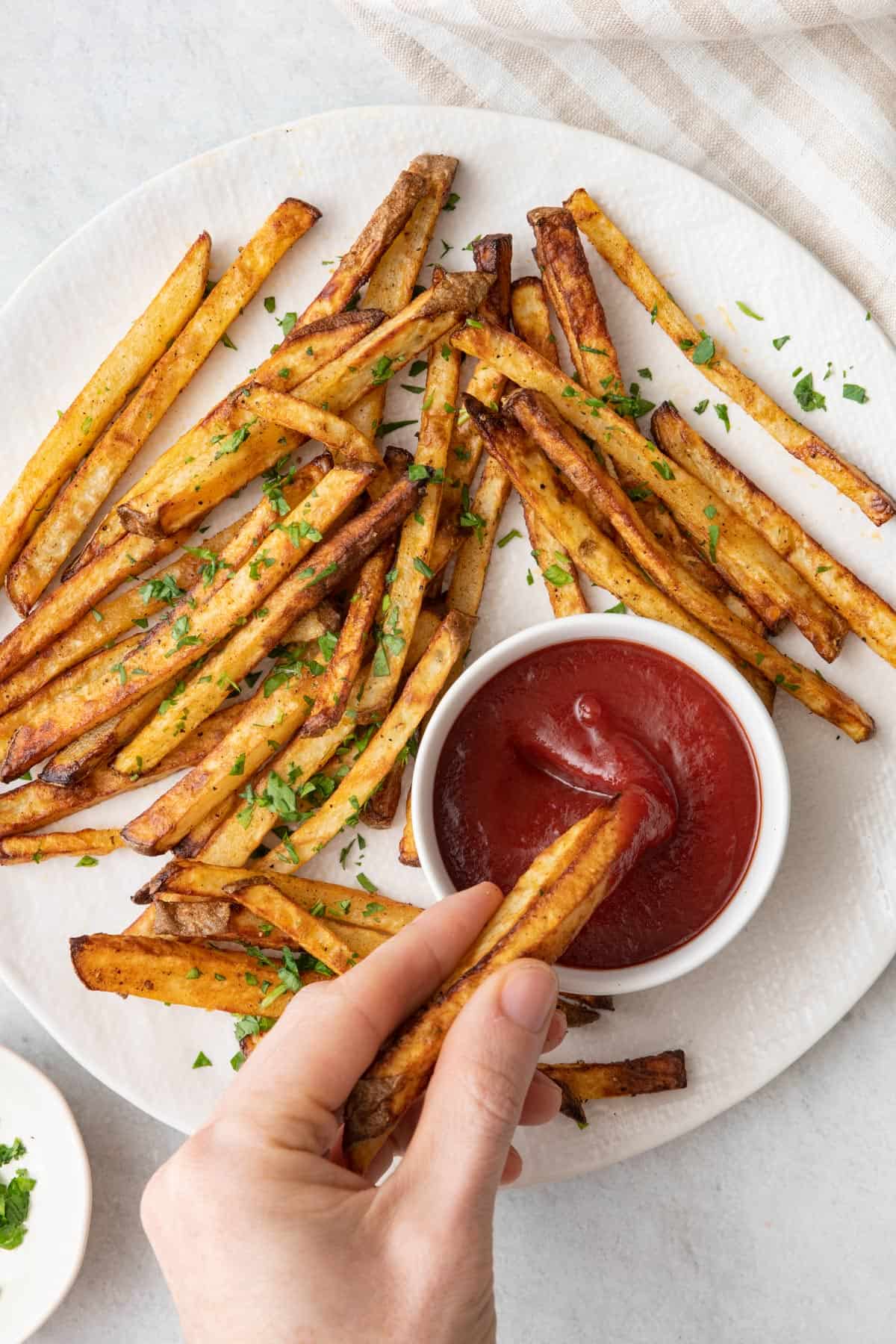 A plate of french fries garnished with parsley with a hand dipping a few into a small dish of ketchup.