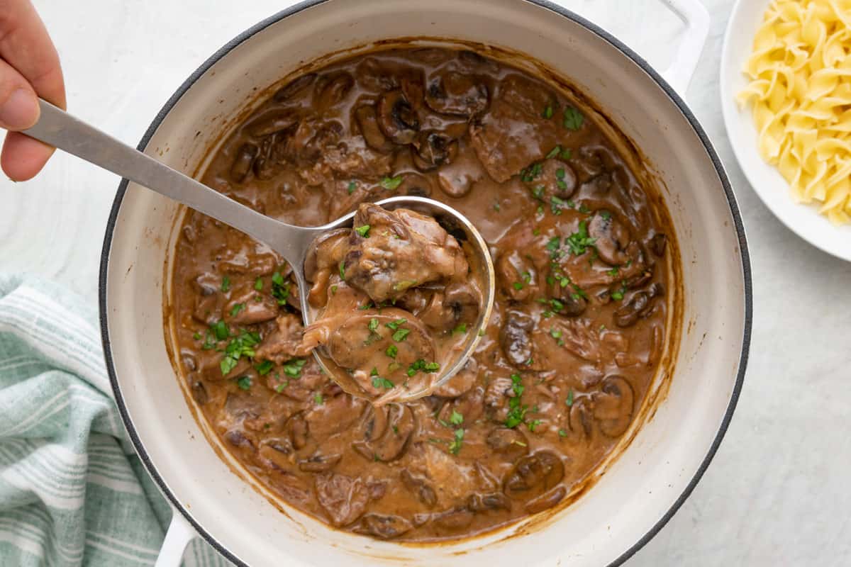 Ladle scooping up some beef stroganoff from the pot to show chunky beef and mushrooms garnished with fresh herbs.