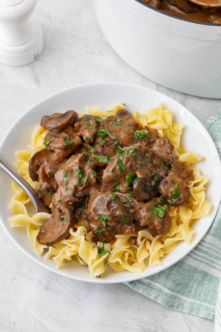 Plate of egg noodles topped with a serving of beef stroganoff, garnished with fresh parsley and a fork dipped inside bowl.