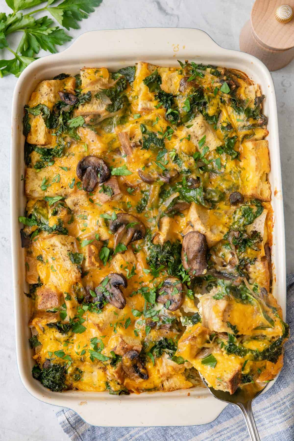 Serving spoon lifting out a serving of cheesy breakfast strata with sliced mushrooms, garnished with fresh herbs.