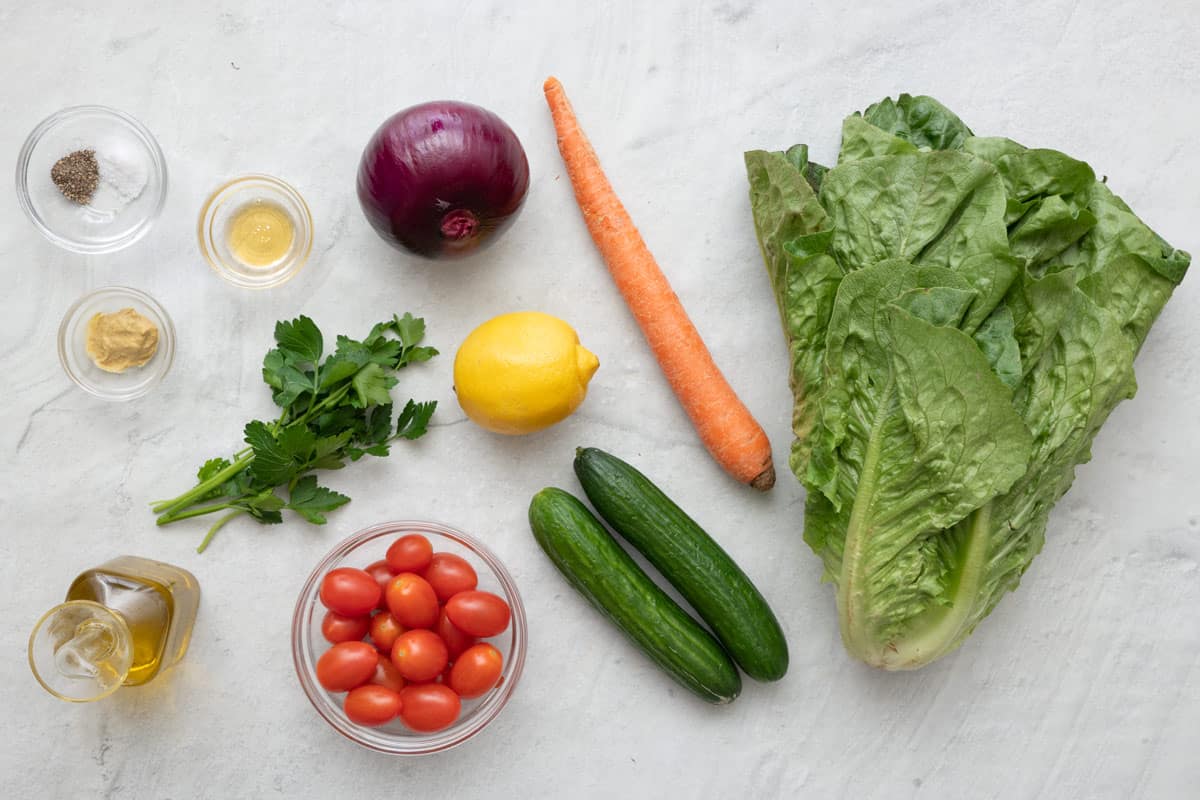 Ingredients for recipe before prepping: seasoning, dijon mustard, oil, honey, parsley, red onion, lemon, persian cucumbers, carrot, and romaine lettuce.