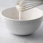 Whisk lifting up icing from a bowl with it falling back into it.