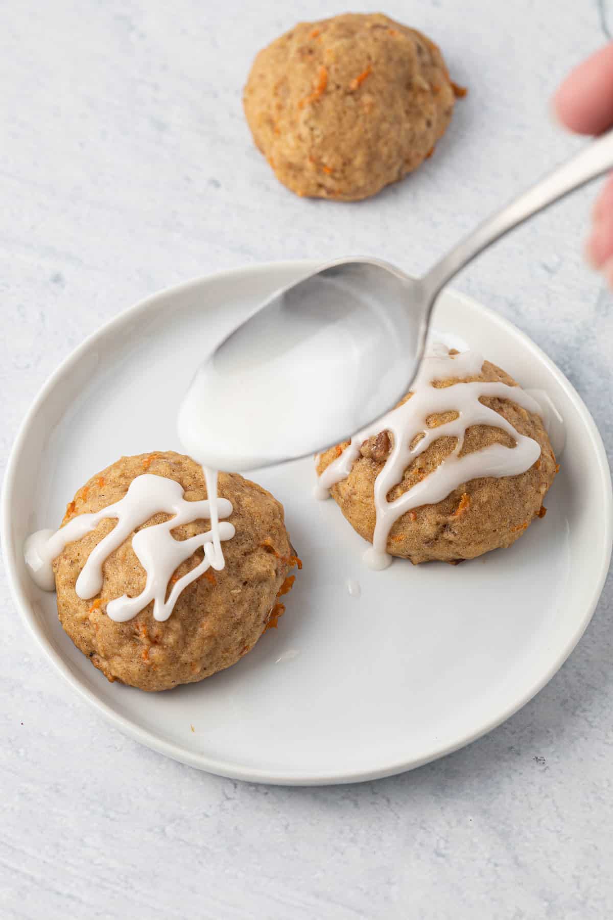 Vanilla icing being drizzled over carrot cake cookies on a plate.