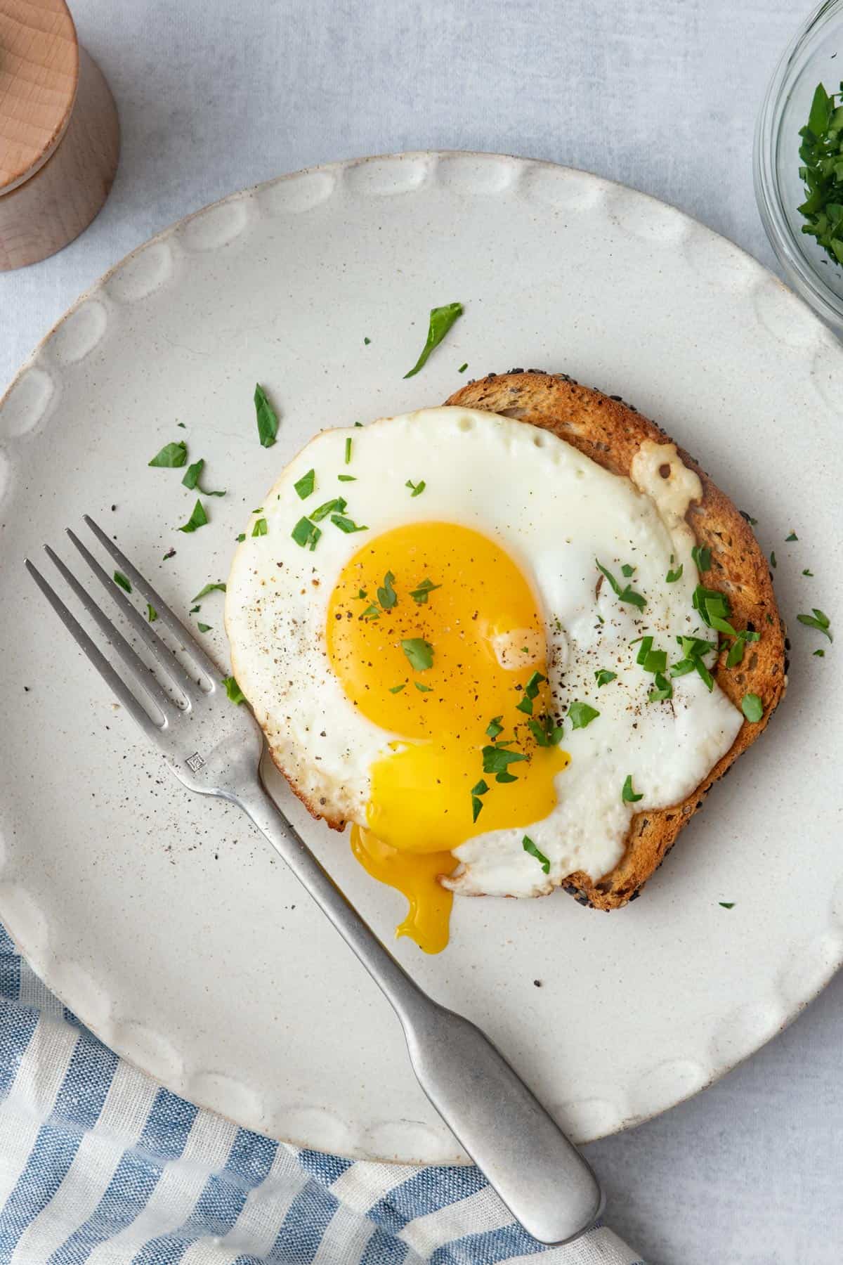 Sunny side up egg over a slice of toast with the yolk broken garnished with fresh herbs on a plate with a fork next to it.