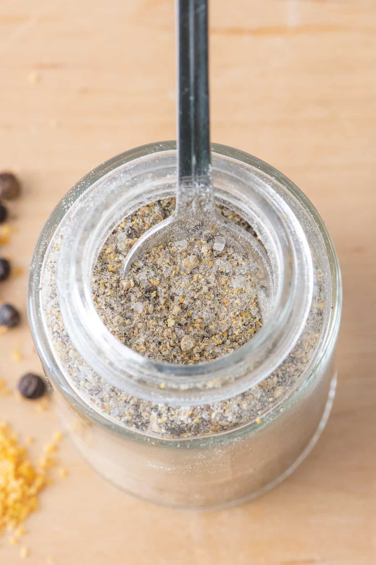 Spice jar filled with lemon pepper seasoning with small baby spoon inside jar to show slightly coarse texture.