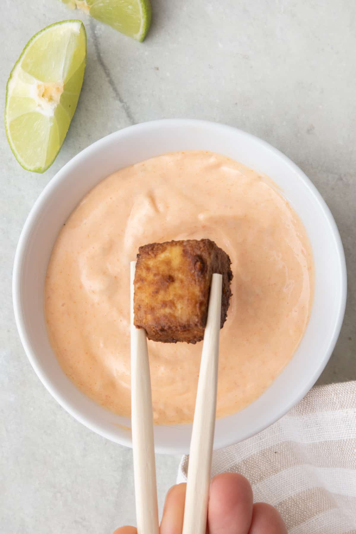 Chopsticks holding a piece of crispy tofu and dipping it into spicy mayo with lime wedges off to the side.