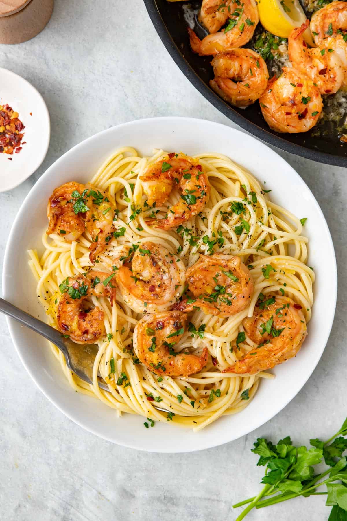 Garlic shrimp served over pasta in a shallow bowl with a forked dipped inside and the pan of shrimp nearby.