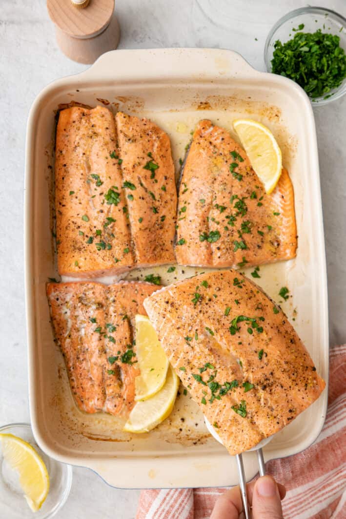Fish spatula lifting up one lemon pepper salmon from baking dish with 3 more fillets garnished with lemon wedges and fresh parsley.