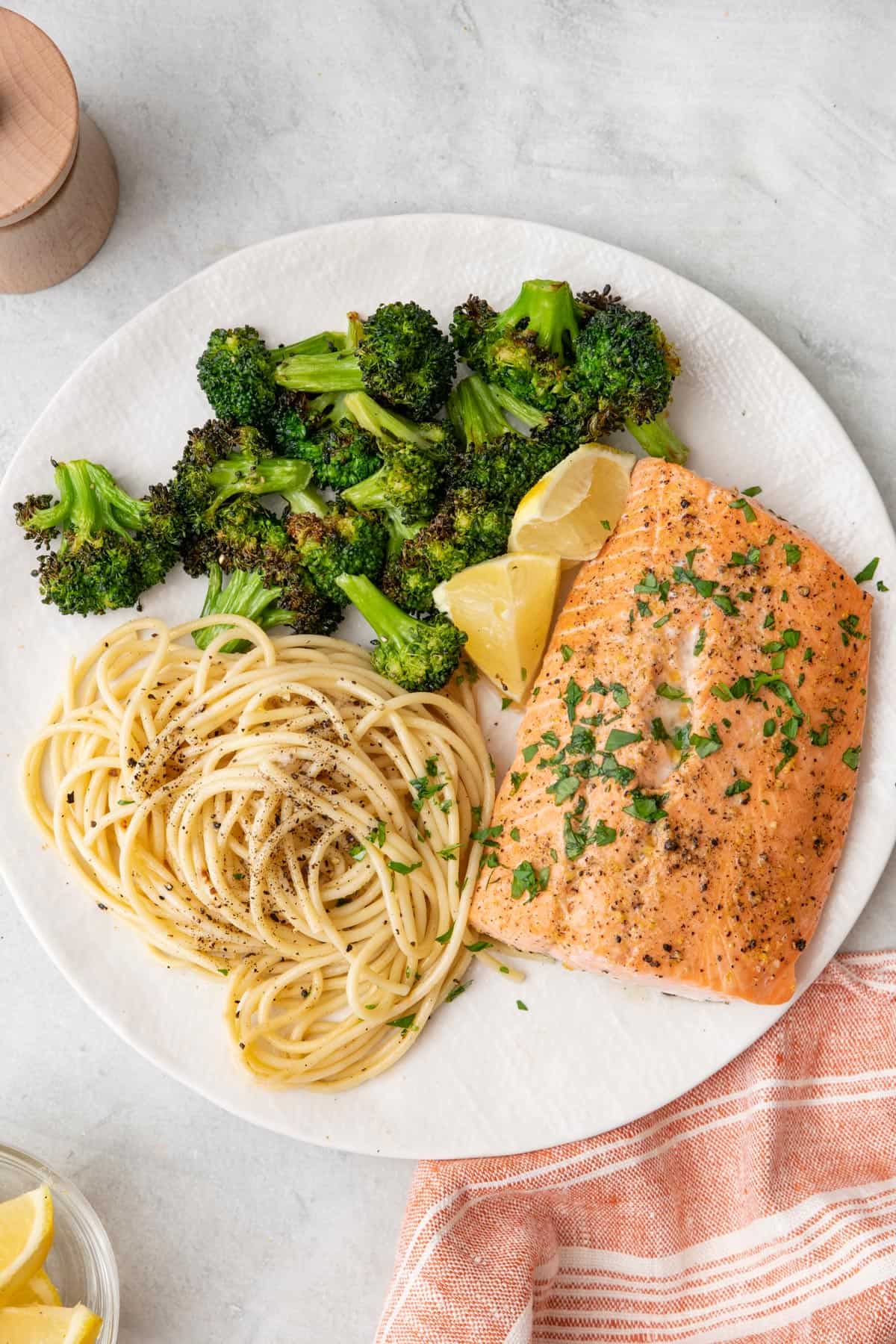 Dinner plate with one salmon filet garnished with fresh parsley and a lemon wedge with a serving of Cacio e Pepe and broccoli