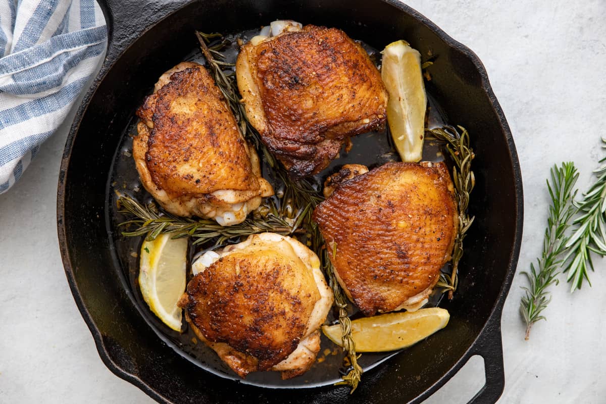 Four seared and roasted chicken thighs with rosemary and lemon wedges baked in a cast iron skillet.