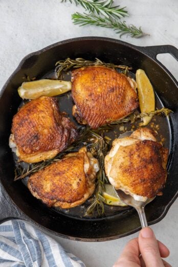 Spatula lifting up a rosemary chicken thigh from skillet with 3 more roasted thighs cooked with rosemary sprigs and garnished with lemon wedges.