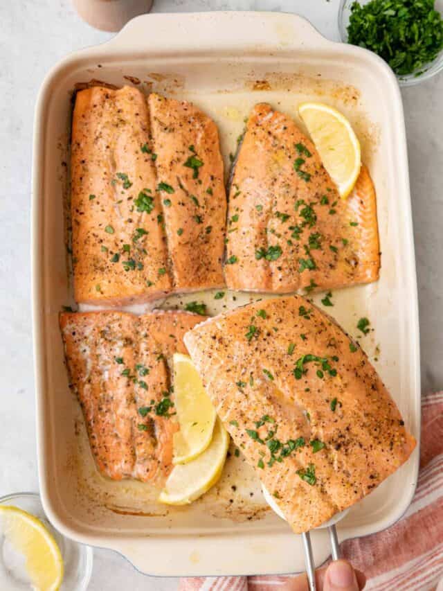 Fish spatula lifting up one lemon pepper salmon from baking dish with 3 more fillets garnished with lemon wedges and fresh parsley.