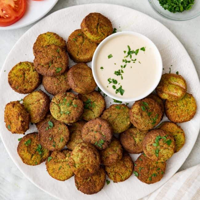 Plate full of air fried falafels with a small dish of tahini sauce and garnished with fresh herbs.