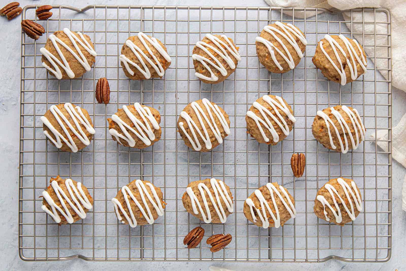 Carrot cake cookies on a wire rack, topped with a drizzle of cream cheese frosting with a few pecans around.