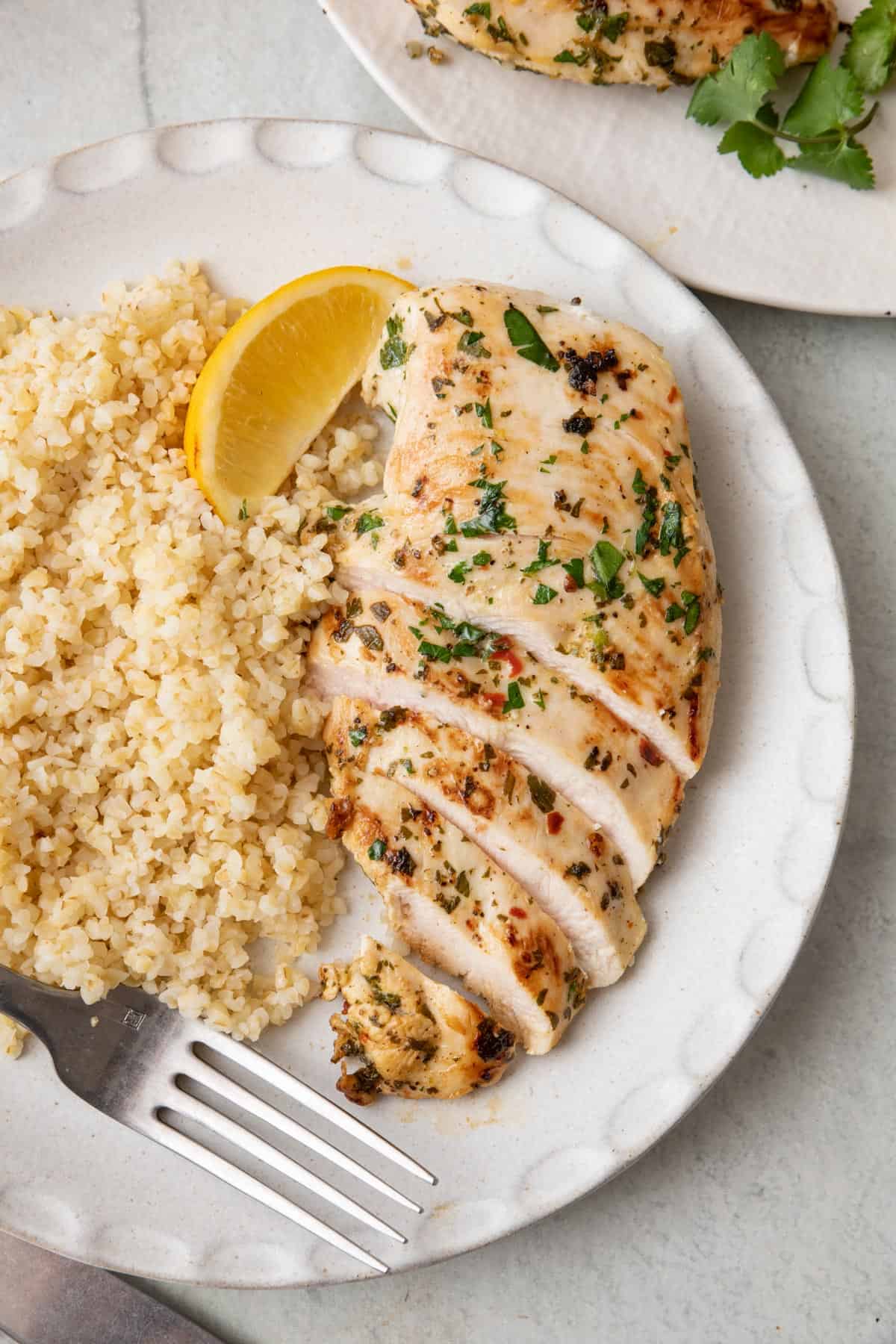 Cilantro chicken on a plate cut into slices halfway through chicken breast with a side of bulgur on a white plate, garnished with a lemon wedge and a fork on plate.