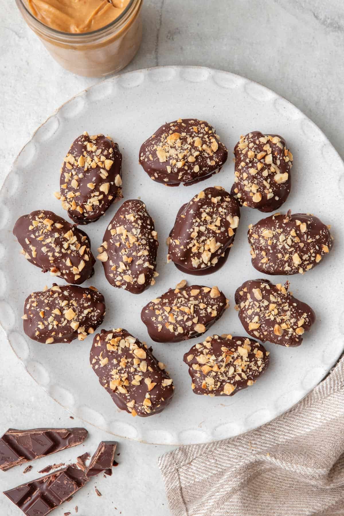 Round white plate with 12 date snickers topped with crushed peanuts with a small jar of peanut butter and chocolate pieces nearby.