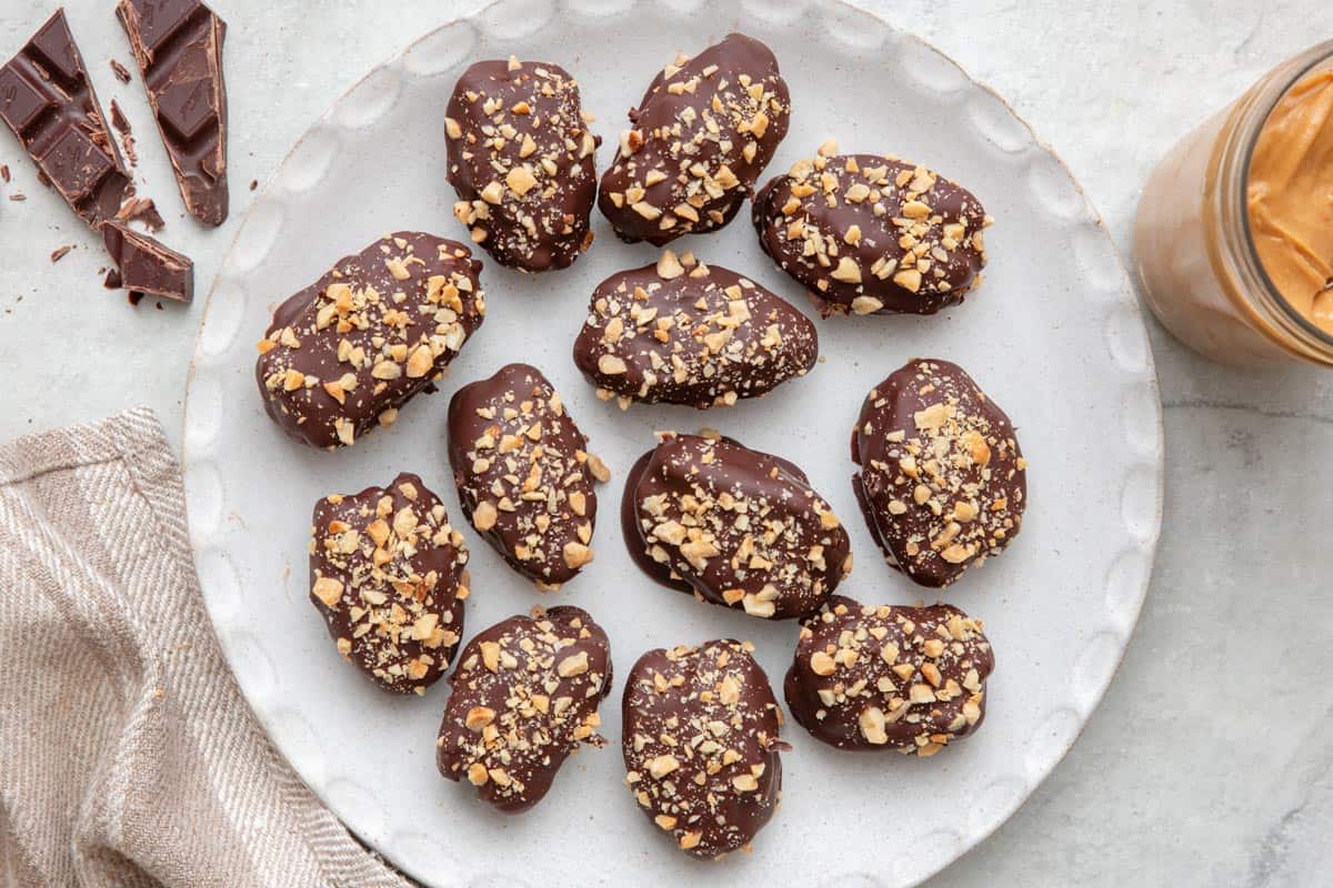 Round white plate with 12 date snickers topped with crushed peanuts with a small jar of peanut butter and chocolate pieces nearby.