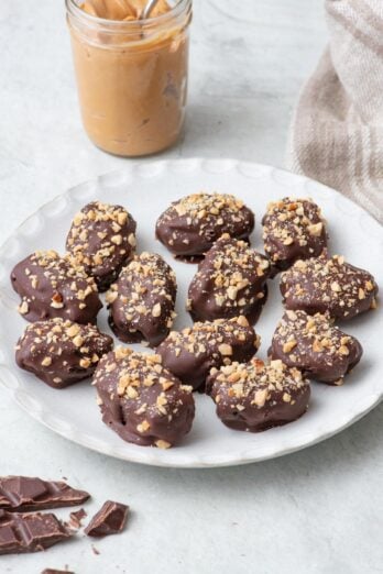 Date Snickers on a round white plate topped with chopped peanuts on top, a small jar of creamy peanut butter with a spoon dipped in and a few chocolate pieces nearby.