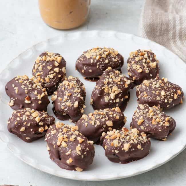 Date Snickers on a round white plate topped with chopped peanuts on top, a small jar of creamy peanut butter with a spoon dipped in and a few chocolate pieces nearby.