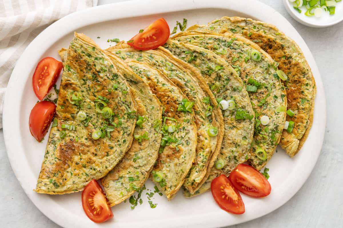 Long round rectangular serving platter with 7 folded omelettes layered across and garnished with green onions, mint and tomatoes.