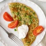 Lebanese omelette on a round plate topped with yogurt, sliced green onions, and chopped mint with a few quartered tomatoes on the side.