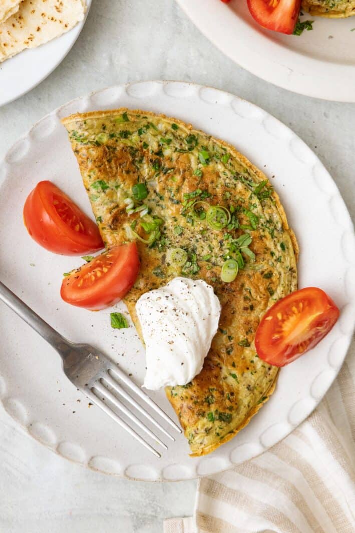 Lebanese omelette on a round plate topped with yogurt, sliced green onions, and chopped mint with a few quartered tomatoes on the side.