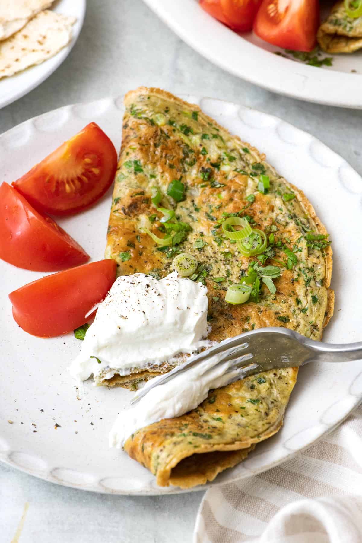 Fork cutting into a Lebanese style omelette called "Ejjeh" topped with yogurt and green onions on a plate with quartered tomatoes.