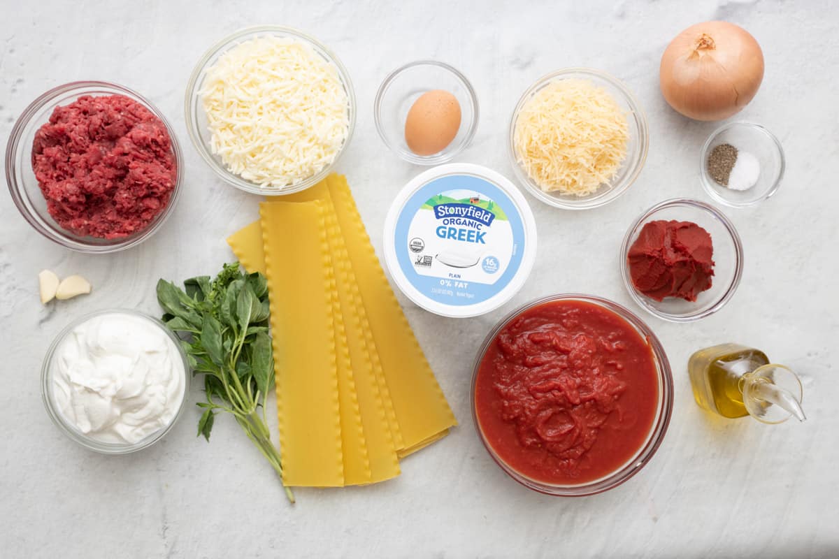Ingredients for recipe: ground beef, garlic cloves, Stonyfield Organic Greek Yogurt in a bowl with container nearby, basil, lasagna noodles, egg, shredded mozzarella, grated parmesan, onion, tomato paste, crushed tomatoes, salt and pepper, and oil.