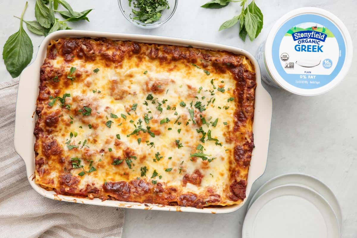 Stonyfield Organic Greek Yogurt Lasagna in a baking dish garnished with fresh basil with 2 small plates nearby and the container for Greek yogurt.