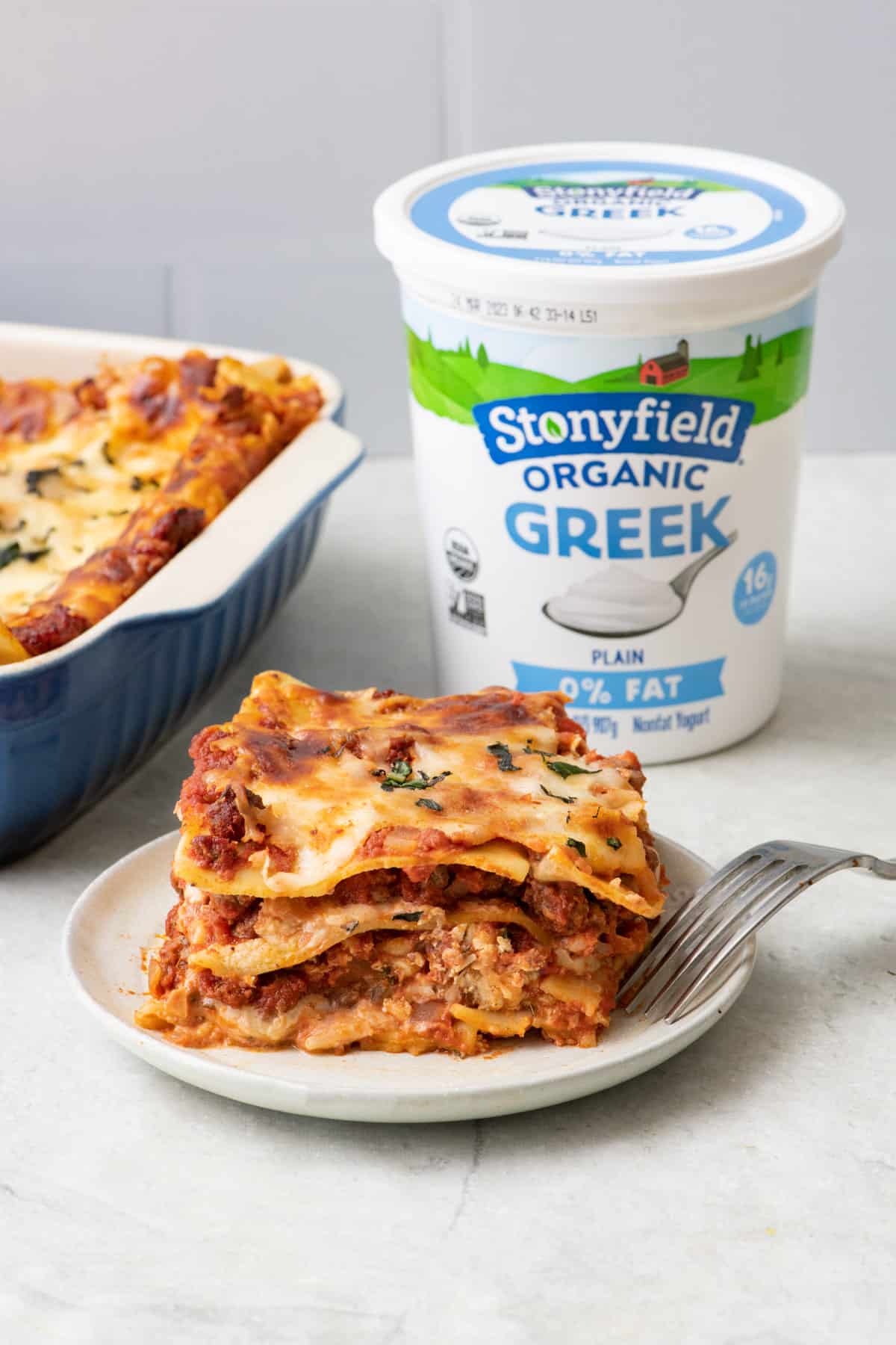 A slice of Stonyfield Organic Greek Yogurt Lasagna on a small plate with baking dish nearby garnished with fresh basil and the container of yogurt behind plate.