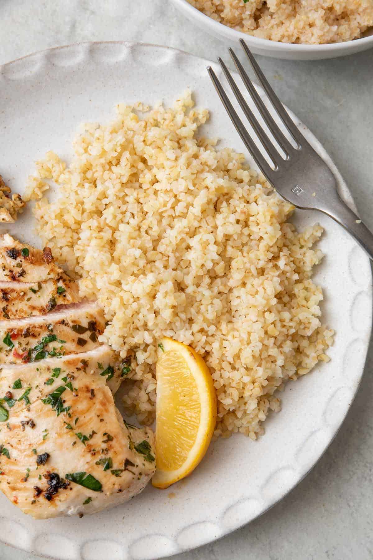 Bulgur served on a plate next to chicken with a lemon wedge and a fork resting on the plate.