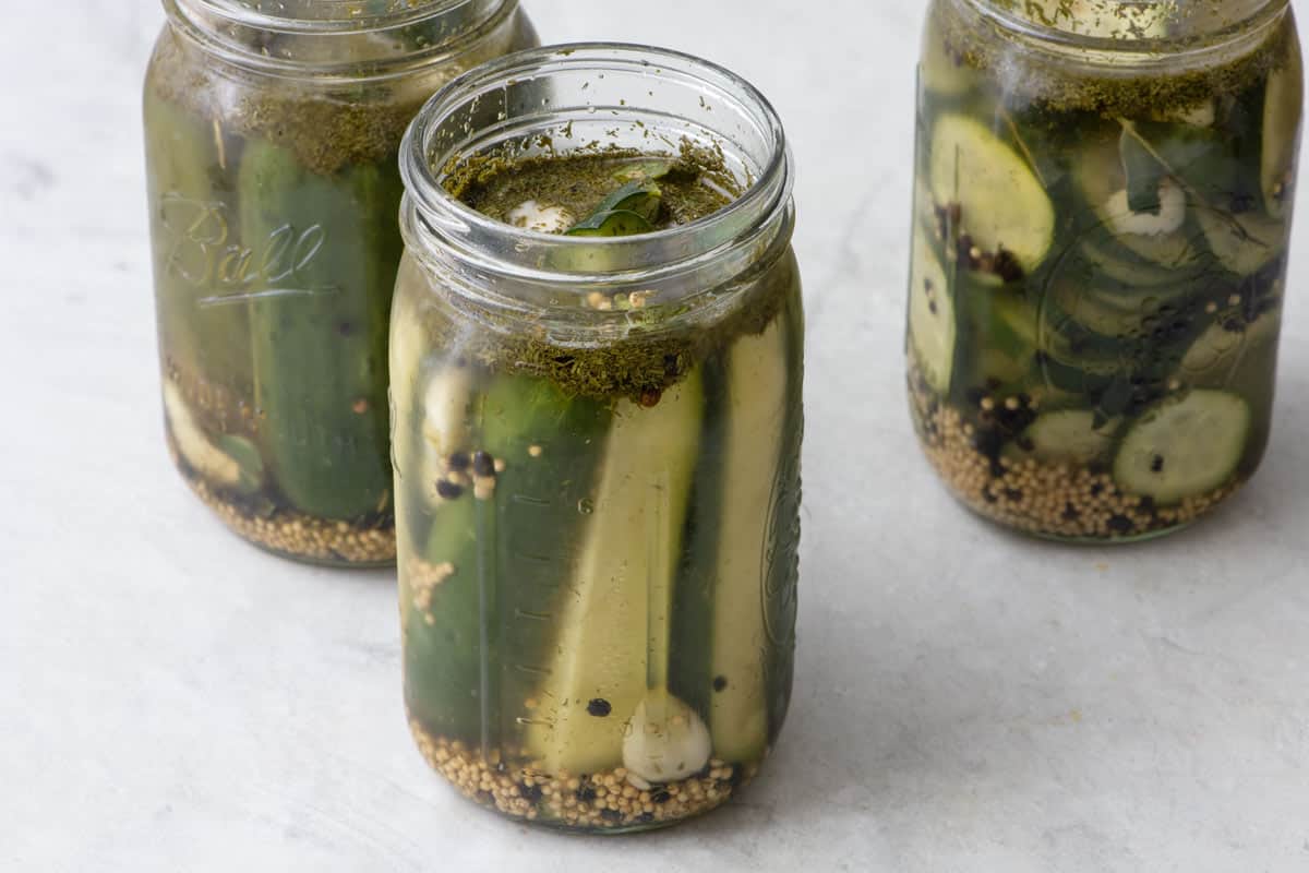 Pickle spears in jar showing seeds and spices floating in brine without lid with with jars of slices and whole pickles in the background.