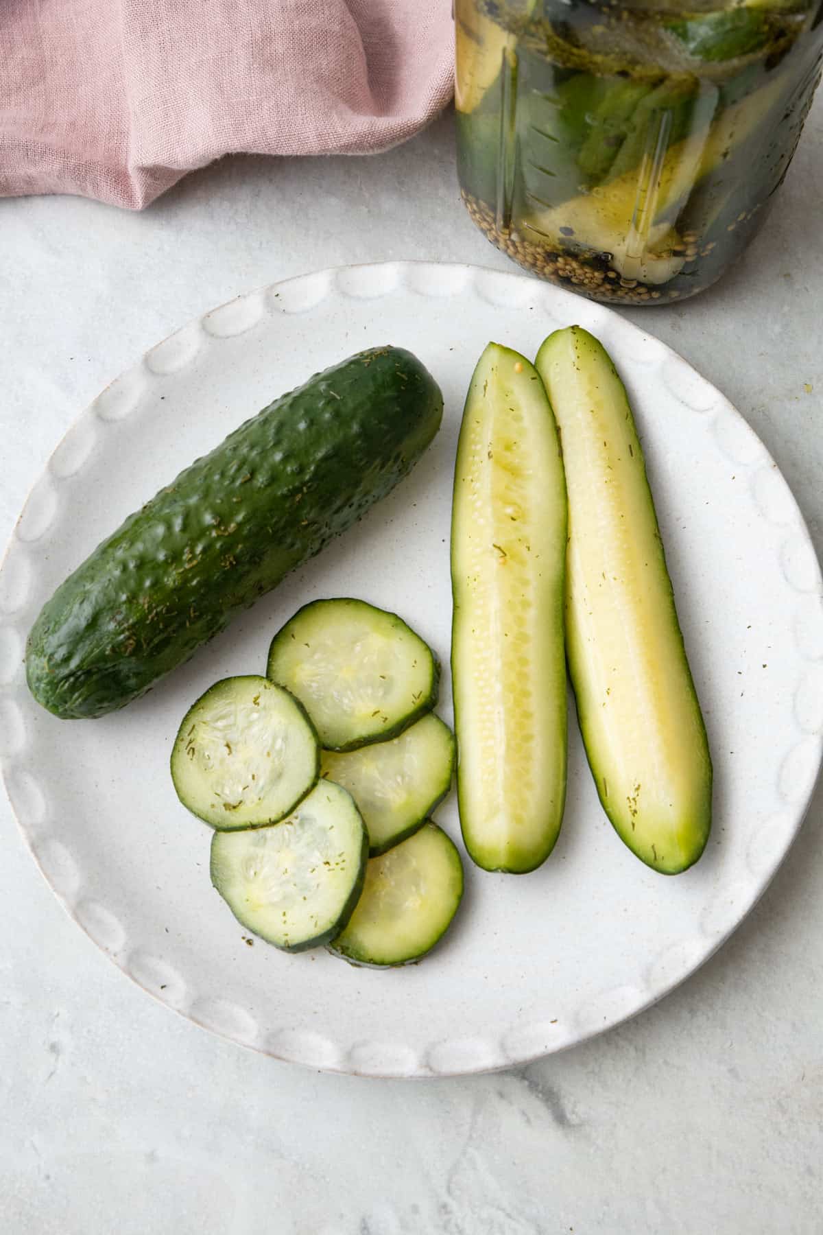 A few pickle spears, slices, and whole pickles on a plate with a pickle jar nearby.