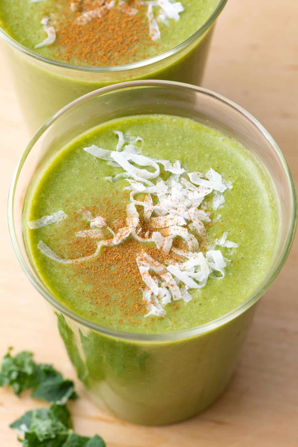 Close up over the top of one glass of kale coconut smoothie to show green color and shredded coconut and cinnamon garnish on top with another glass nearby.