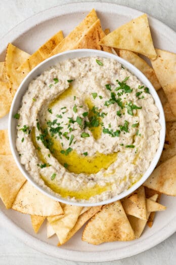 Bowl of baba ghanoush garnished with chopped parsley and olive oil on a platter with pita chips.