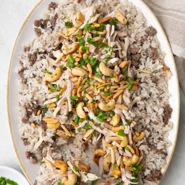 Spoon scooping up a serving of Lebanese chicken and rice topped with toasted nuts on a large oval platter and garnished with fresh parsley.