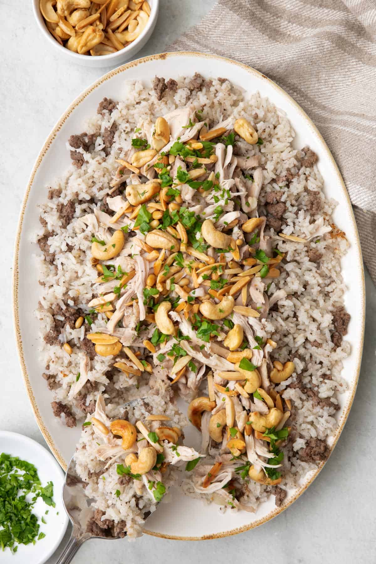 Spoon scooping up a serving of Lebanese chicken and rice topped with toasted nuts on a large oval platter and garnished with fresh parsley.